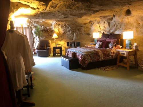 Cozy cave bedroom with a bed, armchair, lamps, and rustic decor, featuring stone walls and soft green carpet.