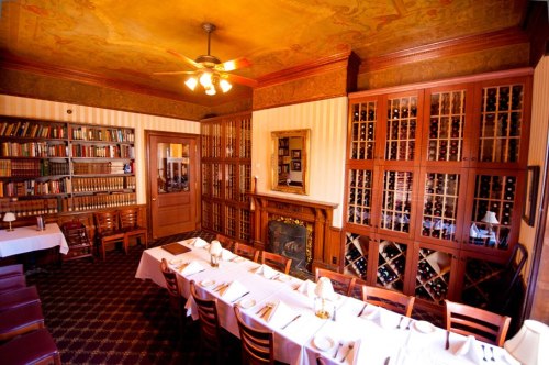 Cozy dining room with a long table, bookshelves, wine storage, and ornate ceiling details. Warm, inviting atmosphere.
