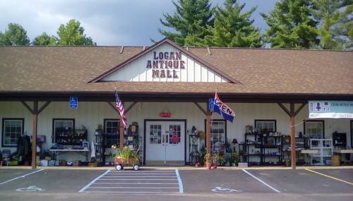 Exterior of Logan Antique Mall with a sign, flags, and various items displayed outside under a cloudy sky.
