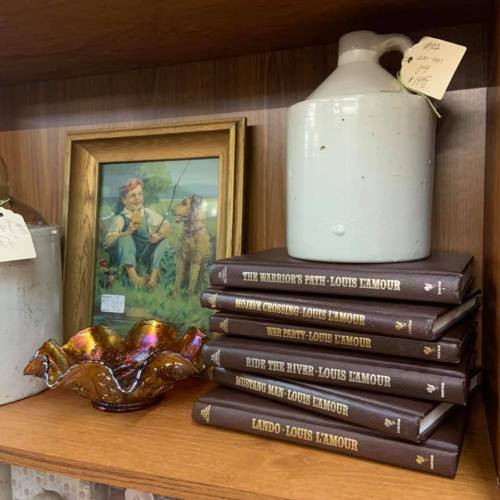 A shelf displaying a vintage jug, framed artwork, and a stack of Louis L'Amour books with a colorful dish.