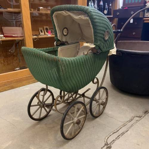 Vintage green wicker baby stroller with metal wheels, displayed in a shop with wooden shelves in the background.