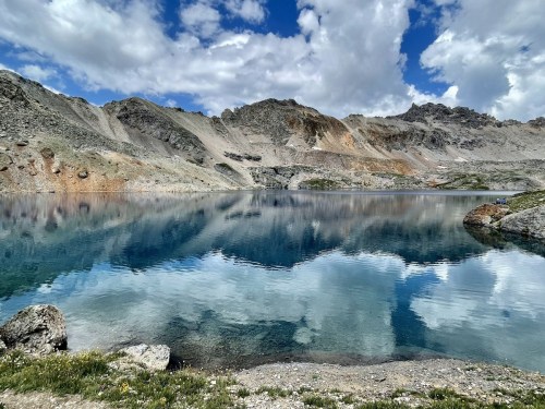 A serene mountain lake reflecting clouds and rocky terrain, surrounded by rugged peaks and greenery.