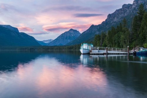 A serene lake at sunset, with a boat docked and mountains reflecting in the calm water under a colorful sky.