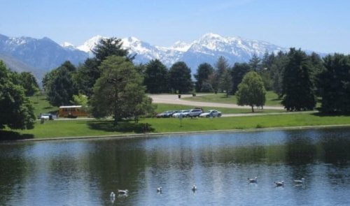 A serene lake reflects snowy mountains, surrounded by lush greenery and a parking area with cars in the background.