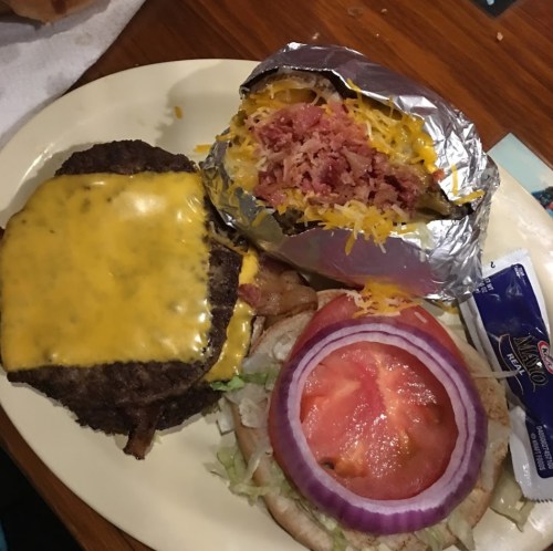 A plate featuring a cheeseburger with melted cheese, lettuce, tomato, onion, and a loaded baked potato wrapped in foil.