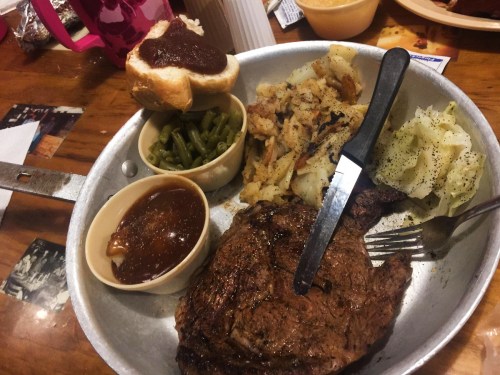 A metal tray with a large steak, green beans, hash browns, cabbage, a roll, and a small bowl of gravy.