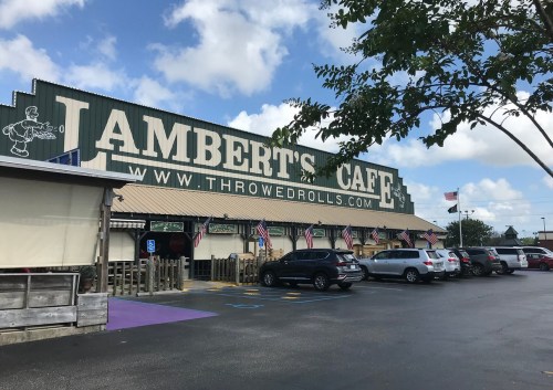 Exterior of Lambert's Cafe with a large sign, parked cars, and American flags under a partly cloudy sky.