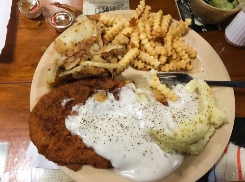 A plate of fried chicken with gravy, mashed potatoes, crinkle-cut fries, and a side of condiments on a wooden table.