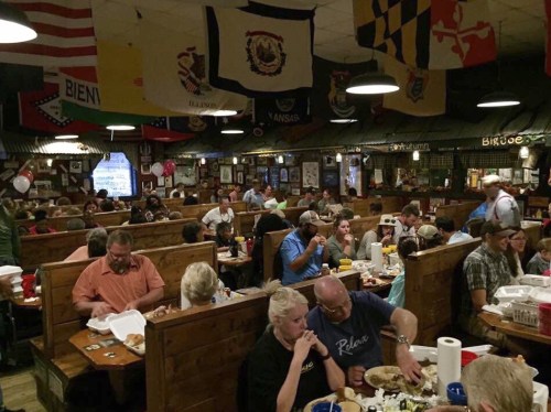 A bustling restaurant filled with diners at wooden tables, surrounded by colorful flags and a lively atmosphere.