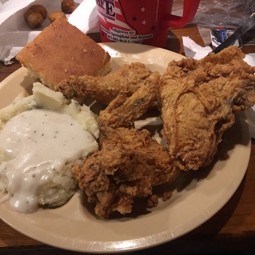 A plate of fried chicken wings, mashed potatoes with gravy, cornbread, and a drink in the background.