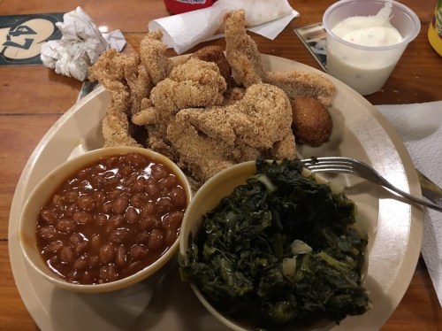 A plate of fried fish, cornbread, baked beans, and collard greens on a wooden table.