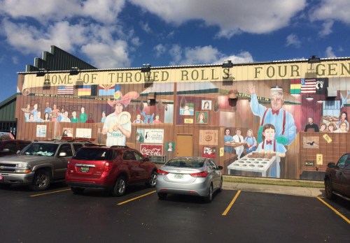 Mural on a restaurant depicting people and scenes related to thrown rolls, with parked cars in front.