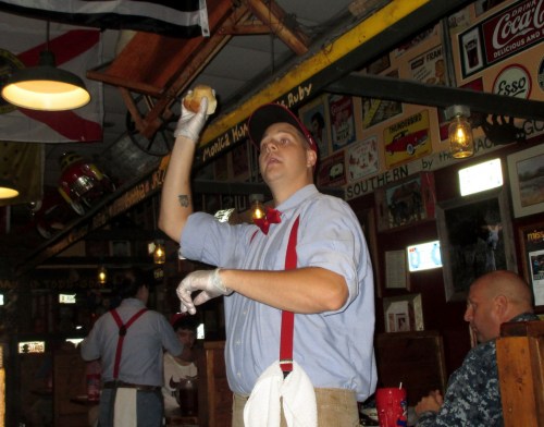 A waiter in a retro diner throws a roll while wearing a red bow tie and suspenders, with vintage decor in the background.