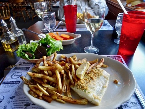 A plate of baked fish with lemon and fries, accompanied by a salad and drinks at a restaurant table.