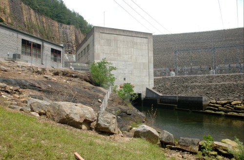A concrete dam structure beside a rocky landscape and a calm water body, with power lines in the background.