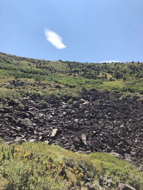 A rocky landscape with green vegetation and a clear blue sky, featuring a single cloud above a sloping hill.