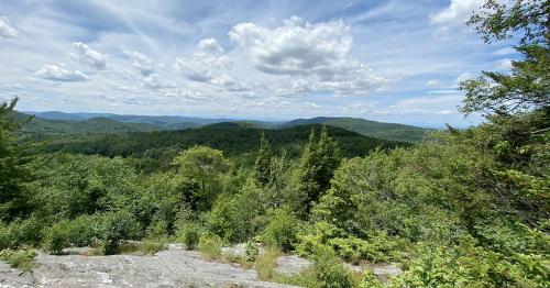 A panoramic view of lush green mountains under a partly cloudy sky, with rocky foreground and distant hills.