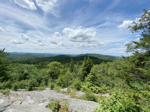 A panoramic view of lush green mountains under a partly cloudy sky, with rocky foreground and distant hills.