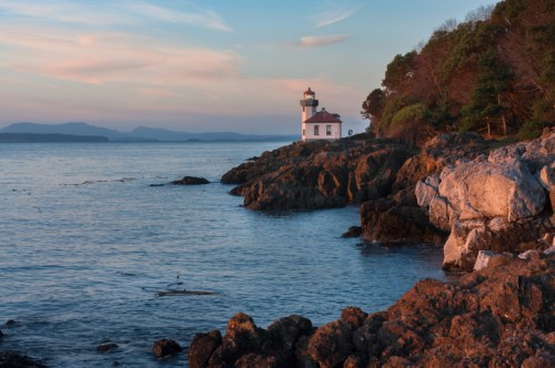 A serene lighthouse stands on rocky shores, surrounded by calm waters and lush greenery under a pastel sky.
