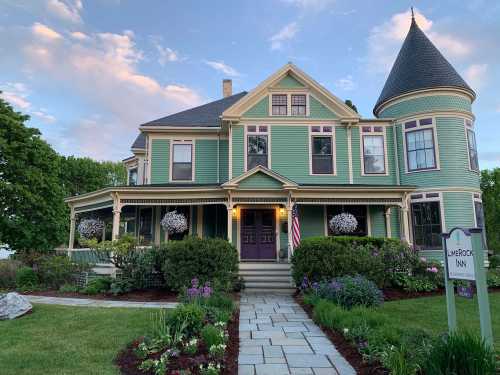 A charming green Victorian inn with a turret, surrounded by lush gardens and flowers, under a blue sky.