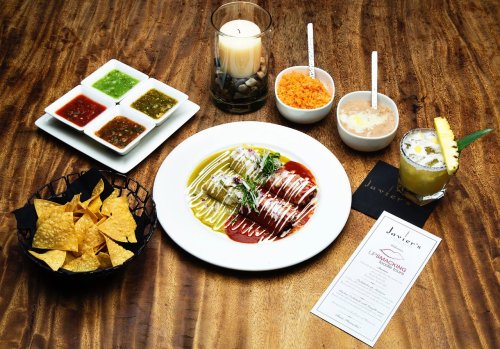 A plate of enchiladas with sauces, accompanied by chips, rice, beans, a candle, and a drink on a wooden table.