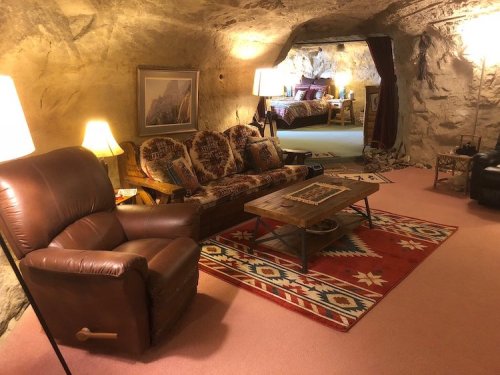 Cozy cave interior with a sofa, armchair, coffee table, and warm lighting, featuring rustic decor and a bedroom in the background.