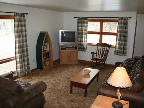 Cozy living room with a TV, rocking chair, and large windows, featuring plaid curtains and a warm color palette.