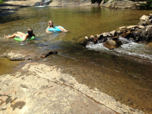 Two people relax on inflatable tubes in a calm, shallow stream surrounded by rocks and greenery.