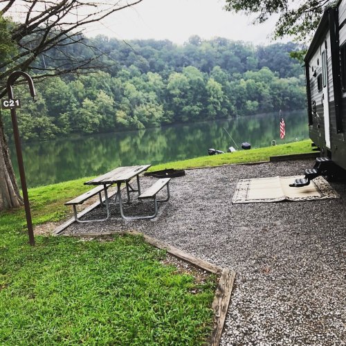 A campsite by a calm lake, featuring a picnic table, a rug, and a camper, surrounded by lush green trees.