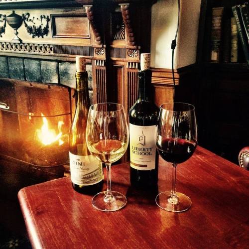 Two wine bottles and three glasses of wine on a wooden table, with a cozy fireplace in the background.