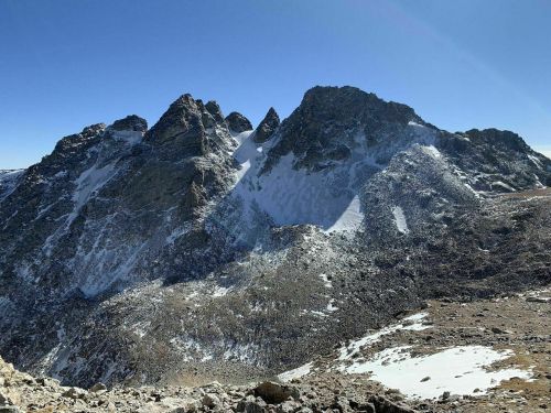 A rugged mountain range under a clear blue sky, showcasing rocky peaks and patches of snow.