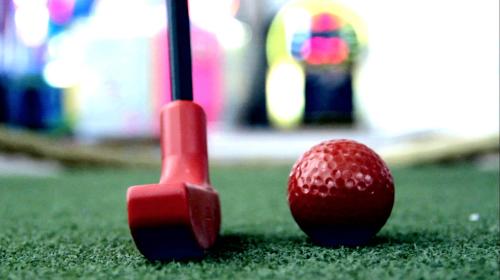 A close-up of a red golf club and a textured red golf ball on green turf, with colorful blurred lights in the background.