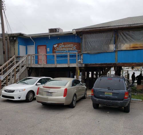 A blue building with a sign reading "Grand Mariner Restaurant & Marina," featuring parked cars and a staircase.