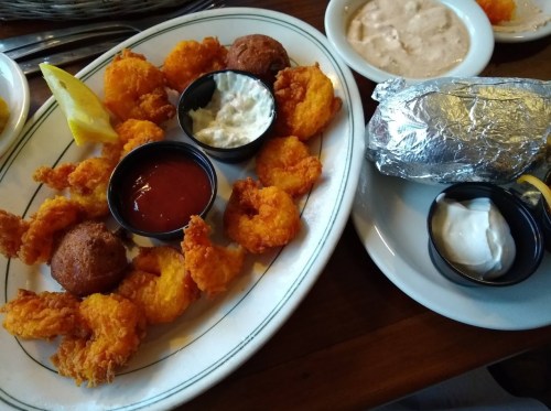 A plate of fried shrimp with dipping sauces, hushpuppies, and a baked potato wrapped in foil.