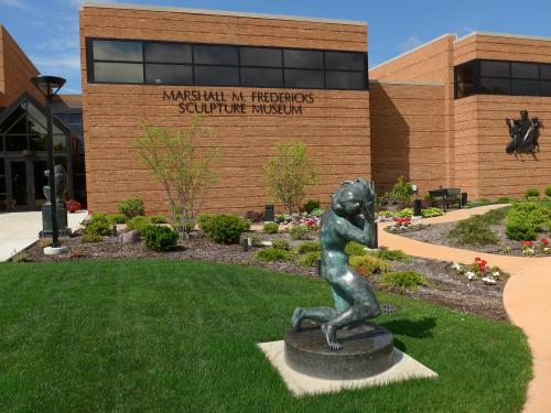 Exterior view of the Marshall M. Fredericks Sculpture Museum, featuring a bronze sculpture in the foreground and landscaped gardens.