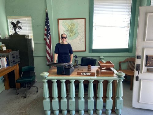 A woman stands behind a vintage sheriff's desk in a green room, with an American flag and a map on the wall.