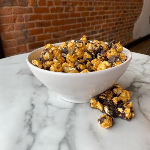 A white bowl filled with caramel and chocolate popcorn, with some pieces scattered on a marble surface.