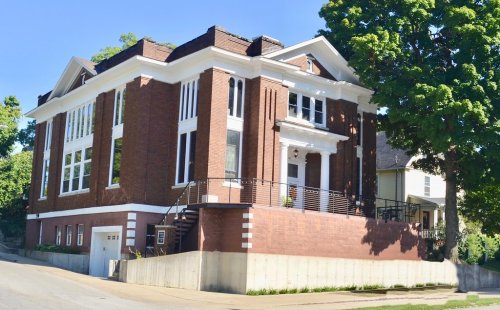 A large, brick building with white trim, featuring multiple windows and a staircase, set on a sloped street.