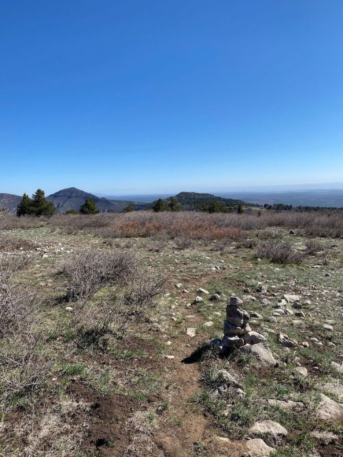 A rocky landscape with a small cairn, rolling hills, and a clear blue sky in the background.