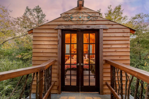 Cozy wooden cabin entrance with double doors, surrounded by greenery and a warm glow from inside.