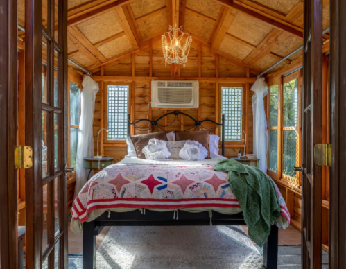 Cozy wooden cabin interior with a bed, chandelier, and air conditioning, featuring large windows and soft decor.