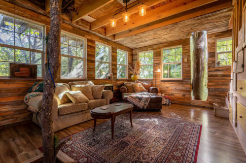 Cozy cabin interior with wooden walls, large windows, a sofa, and warm lighting, creating a welcoming atmosphere.