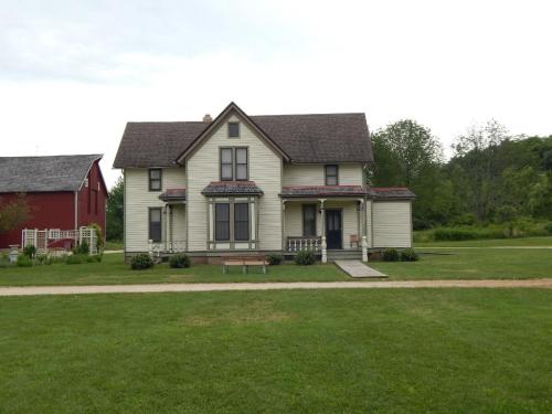 A large, two-story yellow house with a gabled roof, surrounded by green grass and trees, with a red barn in the background.