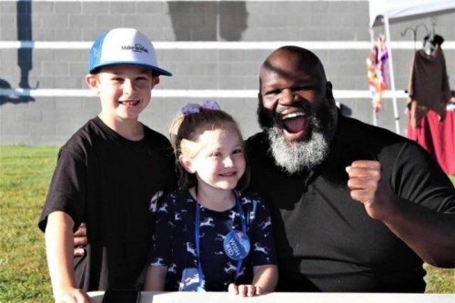 A smiling man with a beard poses with a boy and a girl, both grinning, outdoors at a community event.