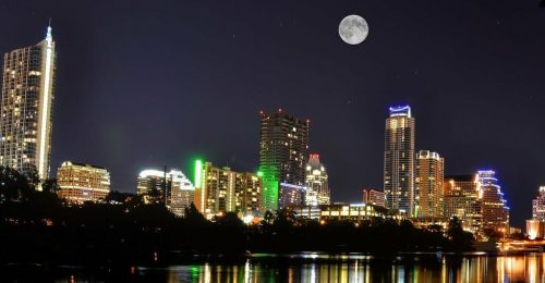 A vibrant city skyline at night, illuminated buildings reflecting on water, with a full moon overhead.
