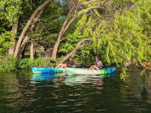 Two people kayaking on a calm river surrounded by lush greenery and trees.