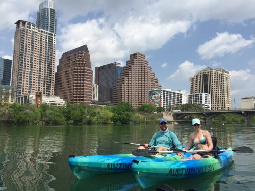 Two people kayaking on a river with a city skyline in the background, featuring tall buildings and greenery.