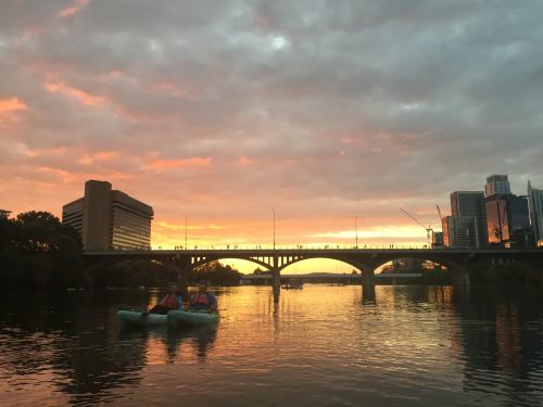 Two kayakers on a river at sunset, with a city skyline and a bridge silhouetted against colorful clouds.