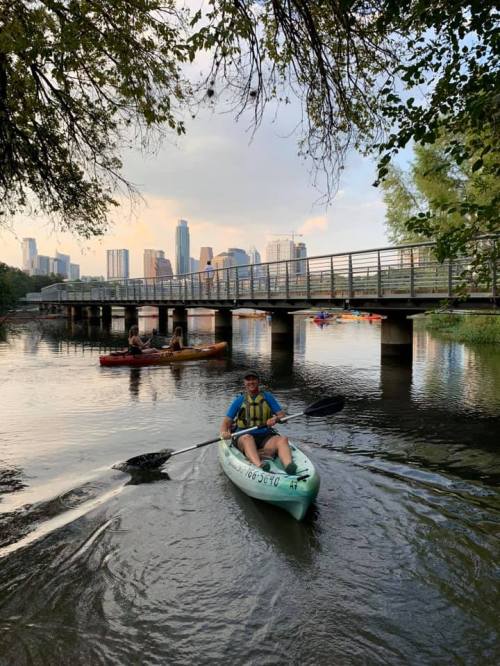 A person kayaking on a river with a city skyline in the background and a bridge overhead. Trees frame the scene.