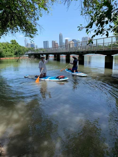 Two people paddleboarding on a calm river with a city skyline and a bridge in the background.